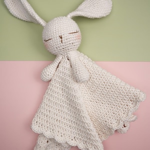 May include: A handmade, off-white crochet bunny lovey with long ears, closed eyes, and a small pink blush on the cheeks. The bunny holds a matching blanket with a scalloped edge, perfect for a baby.