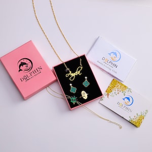 May include: A pink box with a gold necklace, earrings, a ring, and a charm. The necklace has a nameplate that says "Angela". The box has a dolphin logo and the text "Dolphin Jewelry Design". There are two cards with the text "Dolphin Jewelry Design" and "Jewelry Polishing Cloth".