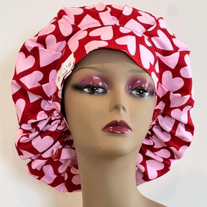 May include: A red and pink heart-patterned bouffant-style hair bonnet. The bonnet features a repeating pattern of pink hearts on a red background. A small tag is visible on the side. The bonnet is designed to cover the hair.
