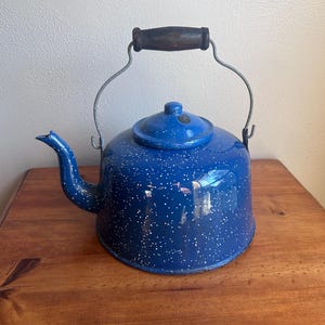 May include: A blue speckled enamelware kettle with a wooden handle and a metal wire frame. The kettle has a spout and a lid. The kettle is sitting on a wooden surface.