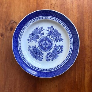 May include: A round, white ceramic plate with a blue floral design. The plate has a blue border with a geometric pattern and a central floral motif. The plate is on a wooden surface.