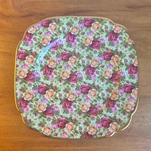 May include: A square ceramic plate with scalloped edges and a gold-trimmed rim. The surface features a dense, repeating pattern of deep red, pink, and yellow roses set against a light green background filled with small green leaves and foliage.