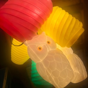 May include: A string of decorative lights featuring translucent plastic owl-shaped covers in white with orange eyes, interspersed with ribbed cylindrical shades in vibrant shades of red, yellow, and green, all illuminated from within.