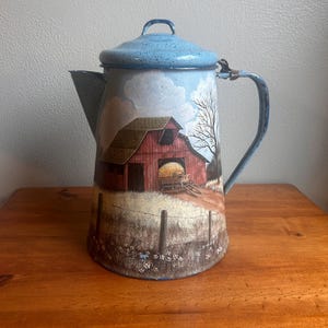 May include: A vintage enamelware coffee pot with a rural landscape scene. The pot features a light blue lid and handle, with a painted image of a red barn, a hay wagon, and a fence. The pot is sitting on a wooden surface.