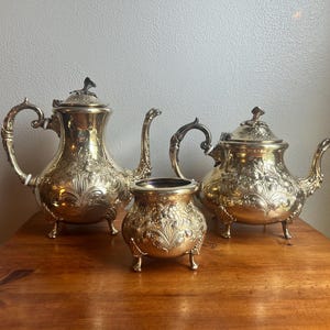 May include: A silver tea set with ornate floral detailing. The set includes a teapot, a creamer, and a sugar bowl, all with matching designs. The pieces are displayed on a wooden surface.