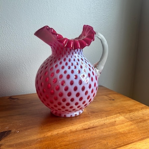 May include: A vintage glass pitcher with a white body and a pattern of red polka dots. The rim and handle are clear glass, and the top edge has a ruffled, deep red trim. The pitcher sits on a wooden surface.
