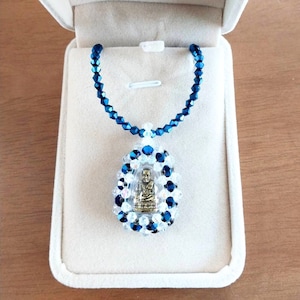 May include: A blue crystal necklace with a clear crystal pendant containing a small gold Buddha statue. The necklace is displayed in a white box.