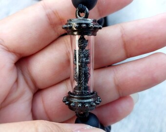 May include: A black beaded necklace with a silver pendant featuring a small statue encased in glass. The pendant has a detailed design with small beads around the top and bottom.