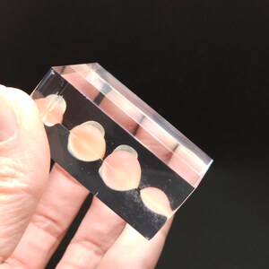 Puede incluir: Un bloque de acr&iacute;lico transparente con cuatro peque&ntilde;os objetos redondos de color rosa claro incrustados en su interior.