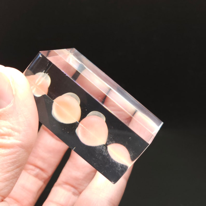 Puede incluir: Un bloque de acr&iacute;lico transparente con cuatro peque&ntilde;os objetos redondos de color rosa claro incrustados en su interior.