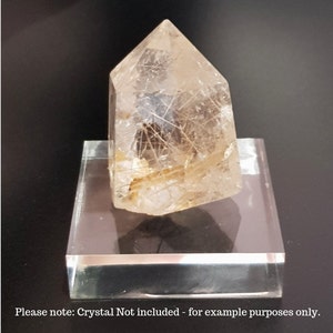 Puede incluir: Un cristal de cuarzo transparente con inclusiones doradas, presentado en un soporte de acr&iacute;lico transparente. El cristal tiene forma de punta y tiene una textura natural y rugosa.