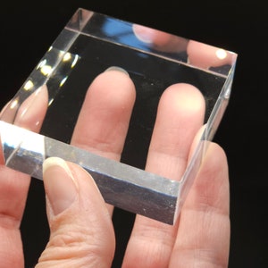 Puede incluir: Un pisapapeles de vidrio transparente con bordes biselados. El pisapapeles es de forma cuadrada y parece estar hecho de acr&iacute;lico.