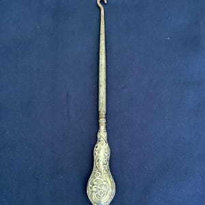 Peut inclure: Un crochet argenté avec un manche décoratif. Le crochet a une extrémité incurvée et une longue tige fine. La poignée présente un motif floral orné. Le crochet est posé sur un fond bleu foncé.