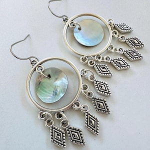 May include: A pair of silver-coloured drop earrings. Each earring has a circular frame with a mother-of-pearl disc and dangling diamond-shaped charms. The iridescent discs reflect shades of blue, green, and silver. The earrings have a hook closure.