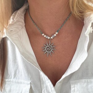 May include: A silver sun pendant necklace with a spiral design and radiating rays. The pendant hangs from a teal cord necklace with silver beads. The necklace is worn over a white shirt.