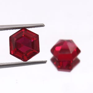 Può includere: Due pietre preziose esagonali rosse, una tenuta da una pinzetta, l'altra su una superficie bianca.