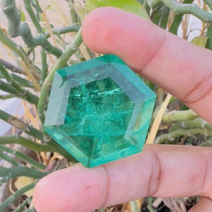 Puede incluir: Una gran gema verde esmeralda facetada con forma hexagonal. La gema se sostiene en una mano, mostrando su tamaño y claridad.