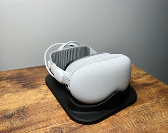 May include: A white virtual reality headset with a black charging stand. The headset is resting on the stand with the cords wrapped around it.