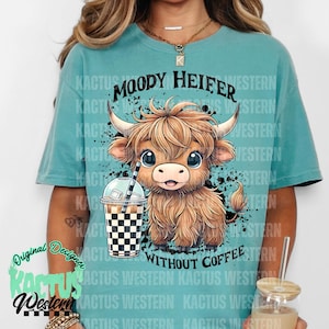 May include: A turquoise blue t-shirt with a black and white graphic of a cartoon Highland cow holding a checkered iced coffee cup. The text "MOODY HEIFER WITHOUT COFFEE" is printed on the shirt.