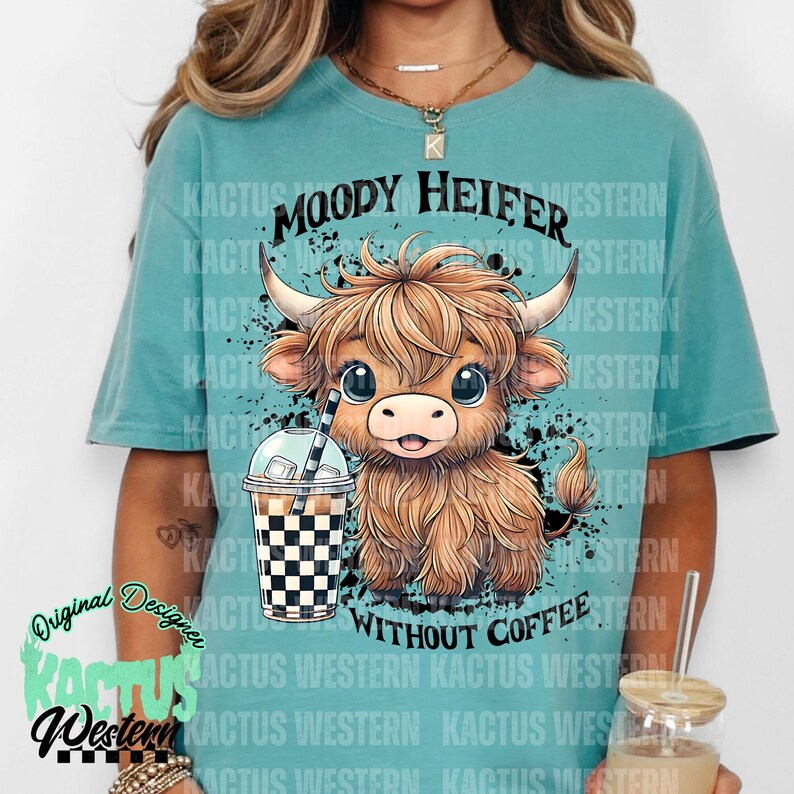 May include: A turquoise blue t-shirt with a black and white graphic of a cartoon Highland cow holding a checkered iced coffee cup. The text "MOODY HEIFER WITHOUT COFFEE" is printed on the shirt.