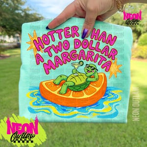 Op de afbeelding: Lichtblauw T-shirt met de roze tekst "Hotter Than A Two Dollar Margarita". Het shirt heeft een cartoon schildpad met een zonnebril, die ontspant op een sinaasappelschijf en een margarita vasthoudt. Het shirt wordt door een persoon omhoog gehouden.