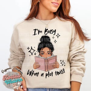 以下が含まれることがあります： 「I'm Busy」と「What a plot twist」の文字が入ったベージュのスウェットシャツ。グラフィックは、本を読む黒髪の女性が特徴です。デザインには星のアクセントと「Kactus Western Co」のロゴが含まれています。