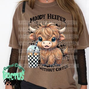 May include: A brown t-shirt with a cartoon illustration of a brown and white highland cow with big eyes and a cup of iced coffee. The text "Moody Heifer Without Coffee" is printed on the shirt.