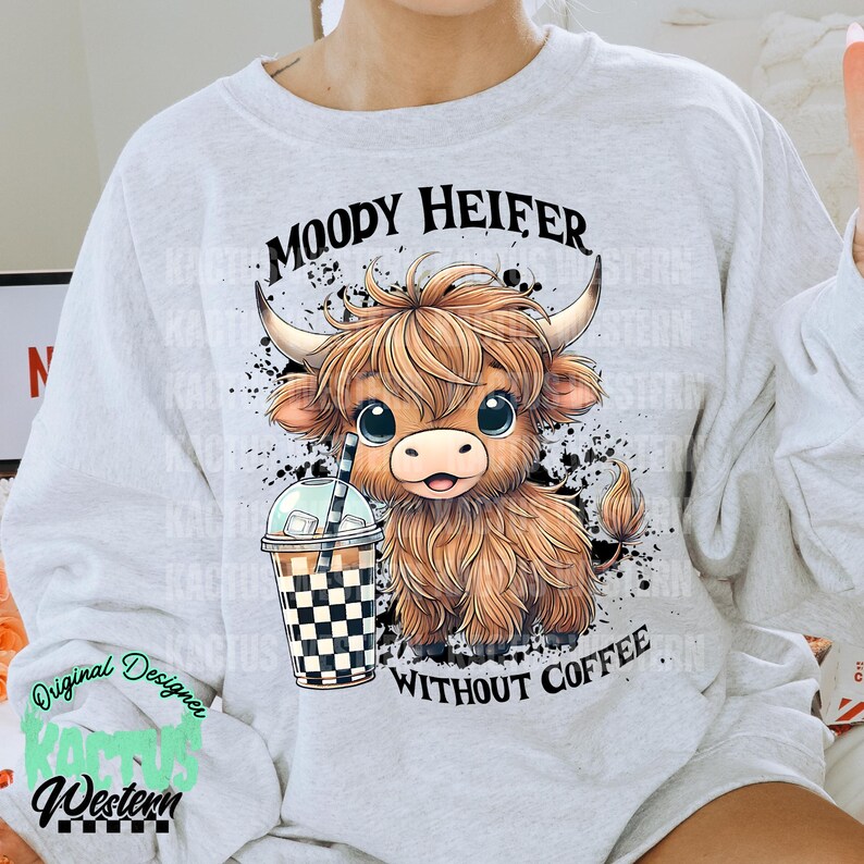 May include: A gray sweatshirt with a cartoon illustration of a brown and white Highland cow holding a checkered cup of coffee. The text "MOODY HEIFER WITHOUT COFFEE" is printed on the sweatshirt.