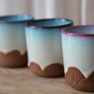 Puede incluir: Tres tazas de cerámica con un degradado de colores. La parte superior de las tazas es azul, desvaneciéndose a blanco y luego a una base marrón. El borde de la taza más a la derecha es de color púrpura intenso. La base de las tazas tiene un diseño de hojas.