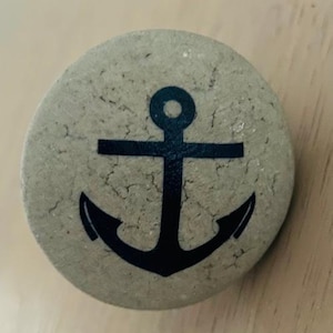 May include: A round, light grey stone coaster with a black anchor design. The anchor has a rounded top and a crossbar.