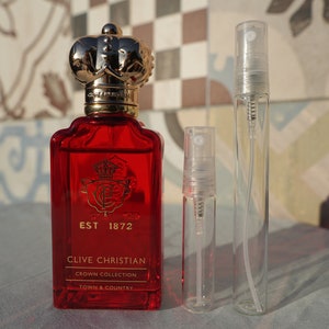 May include: A red glass bottle of Clive Christian Crown Collection Town & Country perfume with a gold crown-shaped stopper. The bottle is next to two clear glass perfume sample bottles with spray tops.