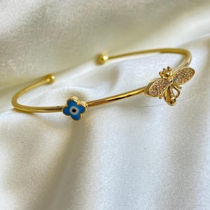 Può includere: Un bracciale rigido color oro con un'ape e un ciondolo a forma di fiore blu. Il ciondolo a forma di ape è impreziosito da piccole pietre trasparenti. Il ciondolo a forma di fiore blu ha un centro bianco e nero. Il bracciale è esposto su un tessuto bianco e setoso.