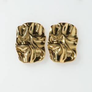 Puede incluir: Un par de pendientes dorados texturizados. Cada pendiente tiene forma rectangular con una superficie irregular y orgánica. Los pendientes son muy reflectantes, creando reflejos y sombras.