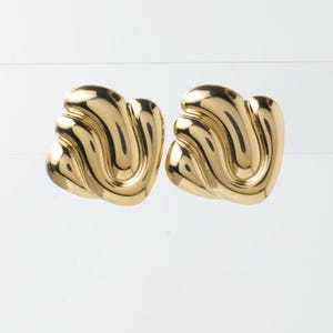 May include: A pair of gold-colored, sculpted earrings. Each earring has a curved, wave-like design. The earrings are made of a shiny, reflective material, and are set against a white background.