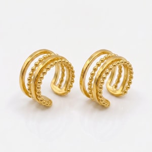 May include: Two gold-colored ear cuffs. Each cuff features three bands, with one band adorned with small, round beads. The other two bands are smooth. The ear cuffs are designed to wrap around the ear.