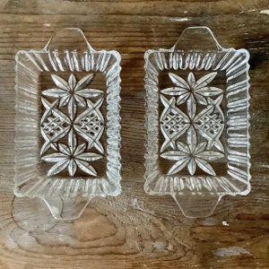 Peut inclure: Deux plateaux rectangulaires en verre transparent avec un motif d'éclat d'étoile et de losange. Chaque plateau a une poignée et un bord texturé et nervuré. Les plateaux sont présentés sur une surface en bois vieilli.