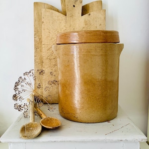 Peut inclure: Un pot en grès vintage avec un couvercle en bois, présenté avec deux cuillères en bois et une planche à découper décorative en bois. Le pot est de couleur marron chaud, et les cuillères et la planche sont brun clair.