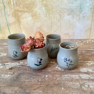 Peut inclure: Quatre vases en céramique faits à la main de formes et tailles variées. Les vases sont de couleur grise mouchetée et décorés de motifs floraux bleus peints à la main. Un vase contient des roses séchées. Les vases sont exposés sur une surface en bois vieilli.