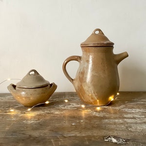 Peut inclure: Deux pièces en céramique : une théière couverte et un petit bol couvert. Les deux sont de couleur beige moucheté avec une finition brillante. La théière a une anse et un bec. Le bol a deux petites anses. Sur une surface en bois rustique.