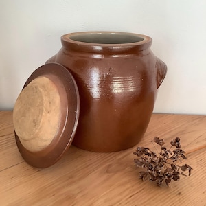 Peut inclure: Un pot en céramique marron avec un couvercle, posé sur une surface en bois. Le pot a une large ouverture et deux petites poignées. Le couvercle est sur le côté, révélant l'intérieur. Un petit arrangement floral séché est au premier plan.