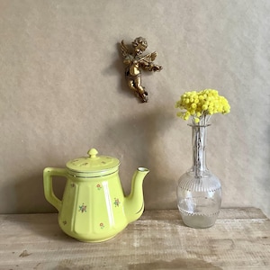 Peut inclure: Une théière jaune avec un couvercle, décorée de petits motifs floraux et de bordures dorées, est posée sur une surface en bois. Un vase en verre transparent contient des fleurs jaunes. Une figurine de chérubin doré est fixée au mur.