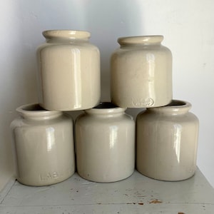 Peut inclure: Cinq pots en céramique blanc cassé, aux corps arrondis et aux bords légèrement évasés, sont empilés sur une surface blanche vieillie. Les pots ont une finition lisse et brillante. Le mot "LAB" est visible sur un pot.