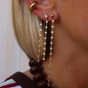 May include: A gold ear cuff, a small stud earring with a clear stone, and a gold ball stud earring, each featuring a long, delicate gold chain that hangs downward. The jewelry is worn on an ear alongside a necklace with large, dark brown beads.