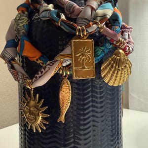May include: A dark blue vase with a textured surface, adorned with a colorful scarf and gold charms. The charms include a sun, a fish, a palm tree, and a seashell. The scarf is multi-colored with various patterns.