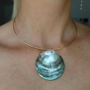 May include: A thin gold-tone metal choker necklace featuring a large, circular, iridescent abalone shell pendant. The shell displays swirling shades of dark green, blue, and grey with a natural pearlescent sheen, suspended from a simple gold wire.