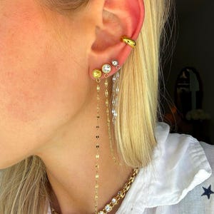 May include: A collection of gold and silver earrings and a necklace. The earrings include a gold ear cuff, stud earrings with yellow and clear stones, and dangling chain earrings. The necklace features a gold chain with clear stones and a gold chain with round gold accents.