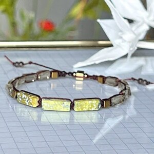 May include: A bracelet made with small, rectangular, light green and gold glass beads. The beads are separated by small, brown, metal beads. The bracelet is tied with a brown cord.
