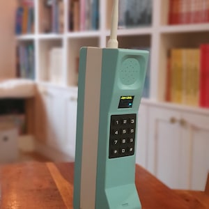 May include: A teal and white retro mobile phone with a long antenna. The phone features a keypad with numbers and symbols, a speaker, and a small screen. The design is reminiscent of early mobile phones.
