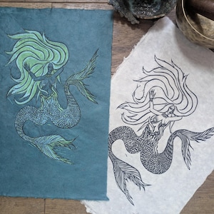 May include: Two mermaid art prints. One print is on teal paper with green and blue details. The other is a black and white print on off-white paper. Both prints feature a mermaid with flowing hair and a fishtail.