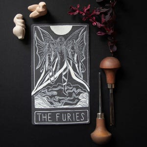 May include: A black and white tarot card print with the text "THE FURIES", depicting three winged figures. The image includes two small sculptures and two wood-handled carving tools. The background is black.